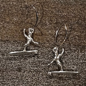 May include: A pair of silver earrings featuring a surfer riding a wave. The earrings are made of metal and have a simple design.