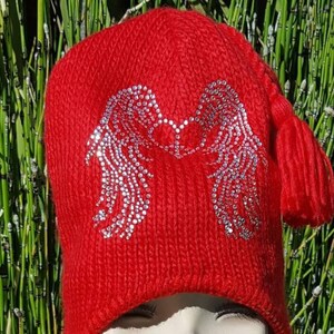 May include: Red knit hat with a large silver sequin design of angel wings. The hat has a long tassel and ear flaps.