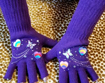 May include: A pair of purple fingerless gloves with colorful heart and butterfly embellishments. The gloves are made of a soft, knitted fabric and have a stretchy fit.
