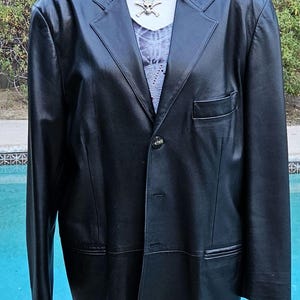 Pu&ograve; includere: Un blazer in pelle nera con revers dentellati, chiusura frontale a un bottone e taschino sul petto. Il blazer &egrave; esposto su un manichino che indossa una maglietta grigia con scollo a V e una collana argentata con ciondolo a teschio.