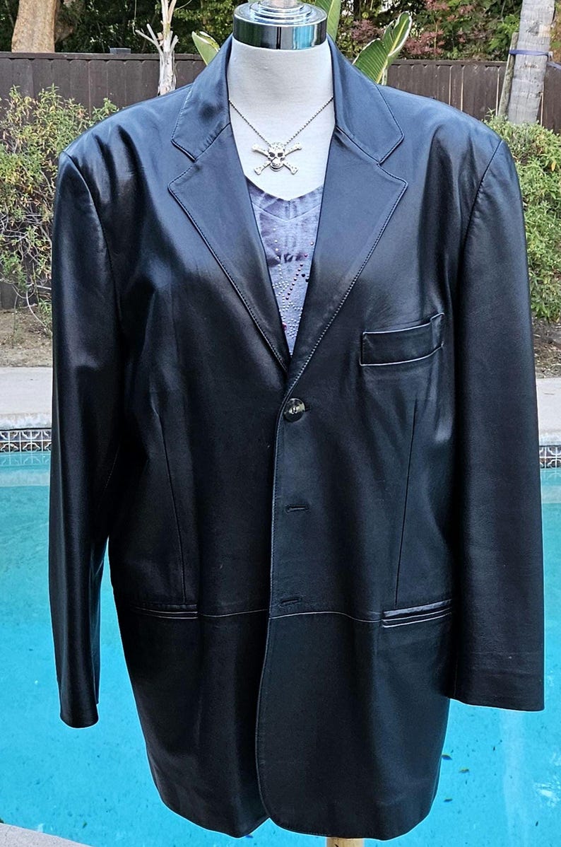 Pu&ograve; includere: Un blazer in pelle nera con revers dentellati, chiusura frontale a un bottone e taschino sul petto. Il blazer &egrave; esposto su un manichino che indossa una maglietta grigia con scollo a V e una collana argentata con ciondolo a teschio.