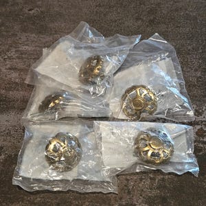 May include: Several decorative cabinet knobs in clear plastic packaging. The knobs have a gold and silver finish with a floral design. The knobs are round and appear to be made of metal. The background is a dark, textured surface.