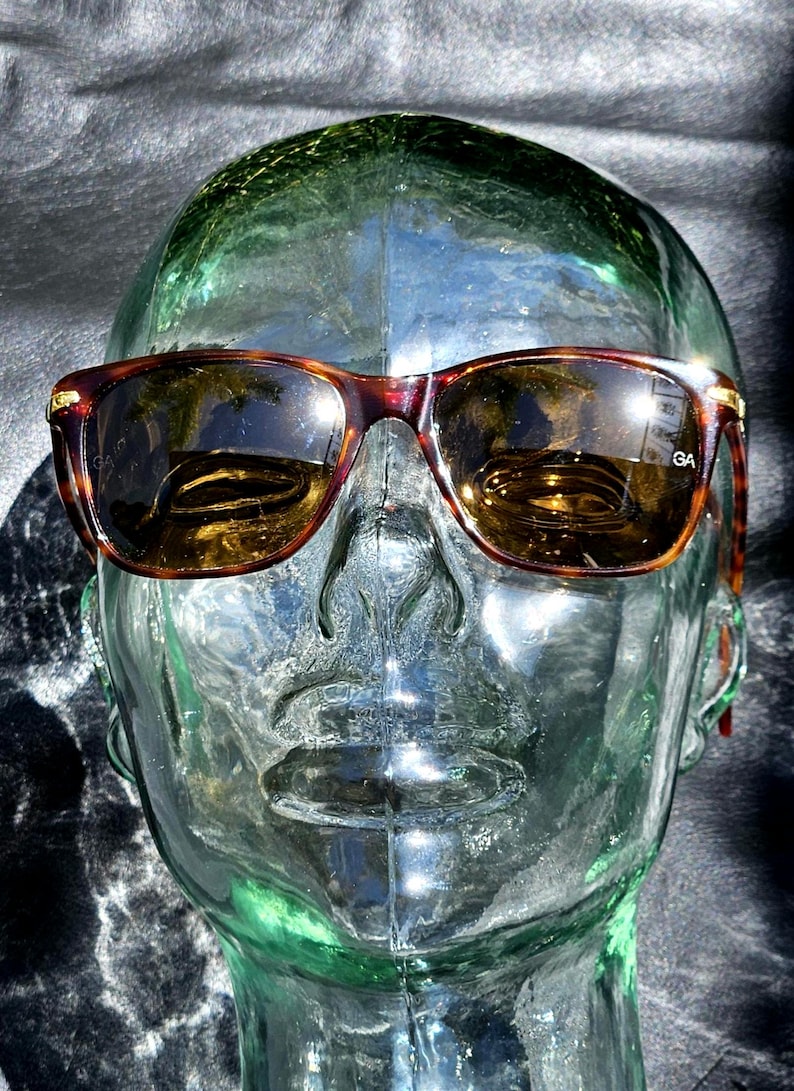 May include: Tortoiseshell-patterned sunglasses with brown-tinted lenses and gold-tone metal accents at the hinges. The right lens features the letters GA printed in white. The frames have a classic rectangular shape with rounded edges.