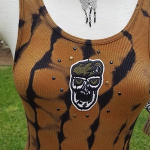 May include: A brown and black tie-dye tank top with a skull and peace sign patch. The tank top has a silver butterfly necklace with black beads hanging from it.