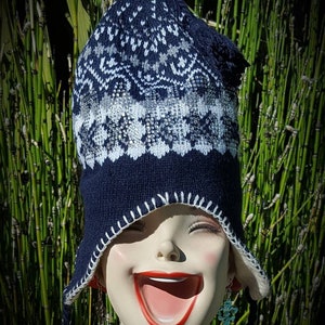 May include: A blue and white knitted hat with a white pom pom on top. The hat has a snowflake pattern and is pulled down over the face of a mannequin.