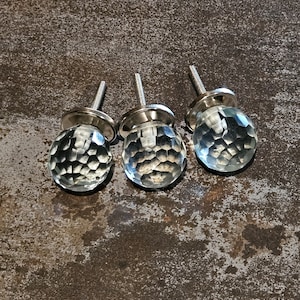 May include: Three clear glass cabinet knobs with a faceted design and silver metal bases. The knobs are arranged in a row on a brown textured surface.