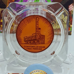May include: A glass ashtray with a red and orange circular sticker featuring a building with the text "Las Vegas Club" and "Downtown Las Vegas Free Parking".  A red dice with white dots and the text "Las Vegas Club" is next to a blue casino chip with a gold $1 and the text "Las Vegas Club Hotel & Casino Las Vegas, NV".