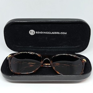 May include: Tortoiseshell-patterned glasses with dark lenses resting inside a black hard-shell case. The interior of the case features the white text READINGGLASSES.COM with a circular logo. The glasses have gold-tone accents on the temples.