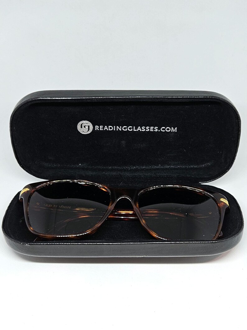 May include: Tortoiseshell-patterned glasses with dark lenses resting inside a black hard-shell case. The interior of the case features the white text READINGGLASSES.COM with a circular logo. The glasses have gold-tone accents on the temples.