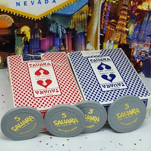 May include: Two red and blue playing card decks with the words "Hotel Sahara Las Vegas, NV" printed on the front. Four gray poker chips with the words "5 Sahara Las Vegas, NV No Cash Value" printed on them.