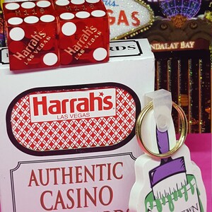 May include: A red box with the Harrah's Las Vegas logo and the text "Authentic Casino Played Cards". Two red dice with the Harrah's Las Vegas logo are in front of the box. A white keychain with a green paint can and the text "Paint the Town Green!" is on the right side of the image.