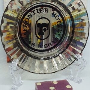 May include: Two purple dice with gold lettering that reads "Frontier Hotel Las Vegas". The dice are sitting on a white surface. A clear glass ashtray with the same lettering is in the background.