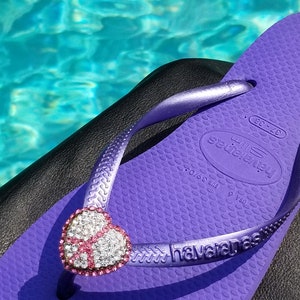 May include: Purple flip-flop with a sparkly pink heart-shaped charm. The flip-flop has the brand name "havaianas" printed on the strap. The flip-flop is on a black surface.