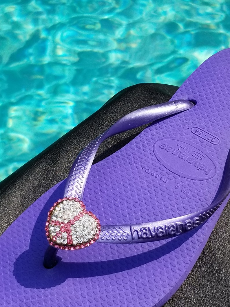 May include: Purple flip-flop with a sparkly pink heart-shaped charm. The flip-flop has the brand name "havaianas" printed on the strap. The flip-flop is on a black surface.