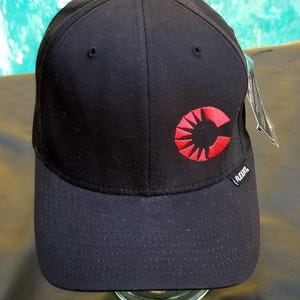 May include: Black baseball cap with a red embroidered logo featuring a stylized letter C with a sunburst design. The logo is centered on the front of the cap. The cap has a curved brim and a black adjustable strap.