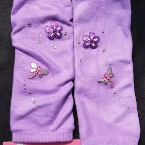 May include: Purple leg warmers with embroidered flowers and dragonflies, embellished with rhinestones. The leg warmers are for infants sizes 6-24 months. The text "infants sizes 6-24m Legwarmers" is printed on a pink label.