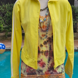 May include: A yellow track jacket with white stripes on the sleeves. The jacket is zipped up and has a tie-dye tank top underneath. The tank top has a lace trim and is worn with a denim skirt.