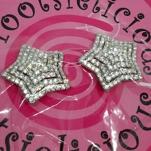 May include: Two silver star-shaped embellishments with clear rhinestones. The stars are on a pink background with the word "lootsielicious" written in black.