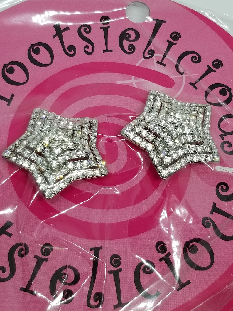 May include: Two silver star-shaped embellishments with clear rhinestones. The stars are on a pink background with the word "lootsielicious" written in black.