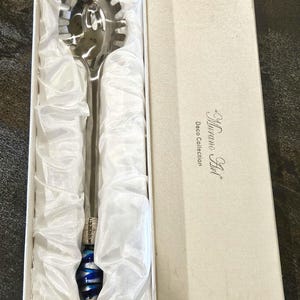 May include: A silver-toned pasta server with a decorative blue and silver handle, presented in a white box. The server has a slotted head for draining pasta. The box is labeled "Murano Art" and "Deco Collection".