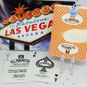 May include: A deck of playing cards with a design featuring the El Rancho Hotel and Casino in Las Vegas, Nevada. The cards are white with black and red suits and a black and white image of the hotel on the back. The box is orange with the El Rancho logo and the words "Hotel & Casino" in white.