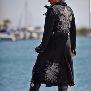 May include: A black long coat with a silver fleur-de-lis design on the back. The coat has a high collar and a belt. The person wearing the coat is standing on a dock with water in the background.