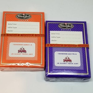 May include: Two packs of Black Jack brand playing cards. The pack on the left is orange with a black and white checkered pattern. The pack on the right is purple with a black and white checkered pattern. Both packs have the text "Black Jack Brand" and "Cartamundi" printed on them. The pack on the left has the text "1SHSH05-22831-A" and "JR GIANT TECH ART" printed on it. The pack on the right has the text "1SHSH06-22175-A" and "JR GIANT TECH ART" printed on it.