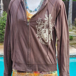 May include: A brown zip-up hoodie with a white crest design on the front. The hoodie has a drawstring hood and a full-length zipper. Layered necklaces and a colorful top are visible underneath. The bottom of the hoodie is visible, along with a denim skirt.