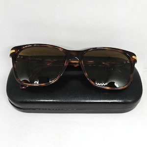 May include: Tortoiseshell-patterned sunglasses with dark brown lenses and gold-tone accents at the hinges. The frames feature the text Giorgio Armani Made Italy on the inner temple. The sunglasses are resting on a black rectangular hard case.