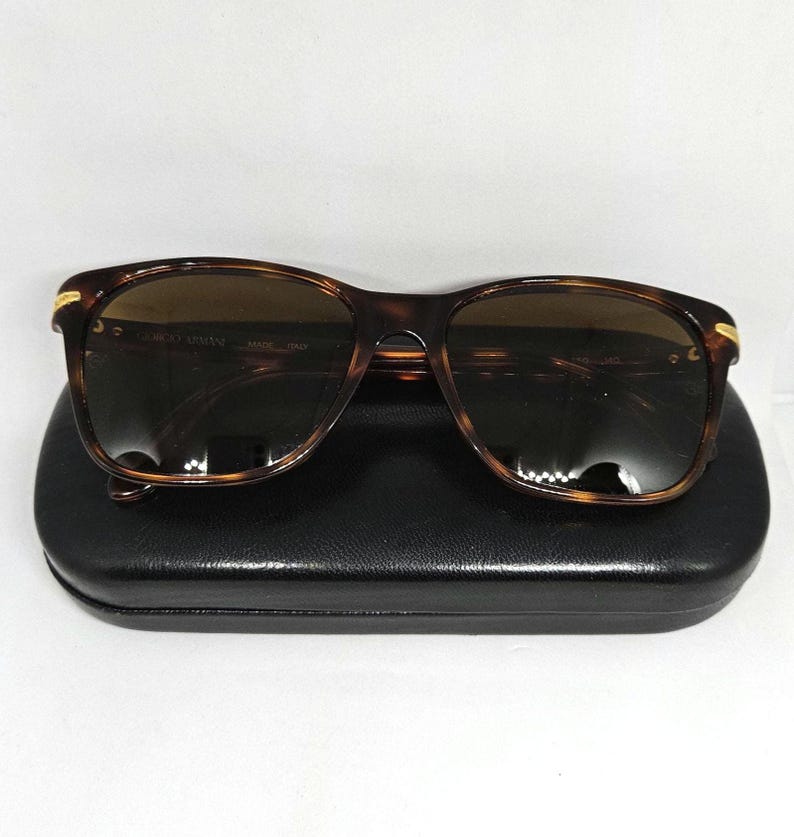 May include: Tortoiseshell-patterned sunglasses with dark brown lenses and gold-tone accents at the hinges. The frames feature the text Giorgio Armani Made Italy on the inner temple. The sunglasses are resting on a black rectangular hard case.