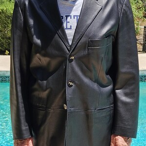 Pu&ograve; includere: Un blazer in pelle nera con revers dentellato, tre bottoni frontali e una tasca sul petto. Il blazer &egrave; indossato sopra una t-shirt grigia con il testo "ET" visibile in lettere blu. Il capo presenta maniche lunghe e un orlo dritto.