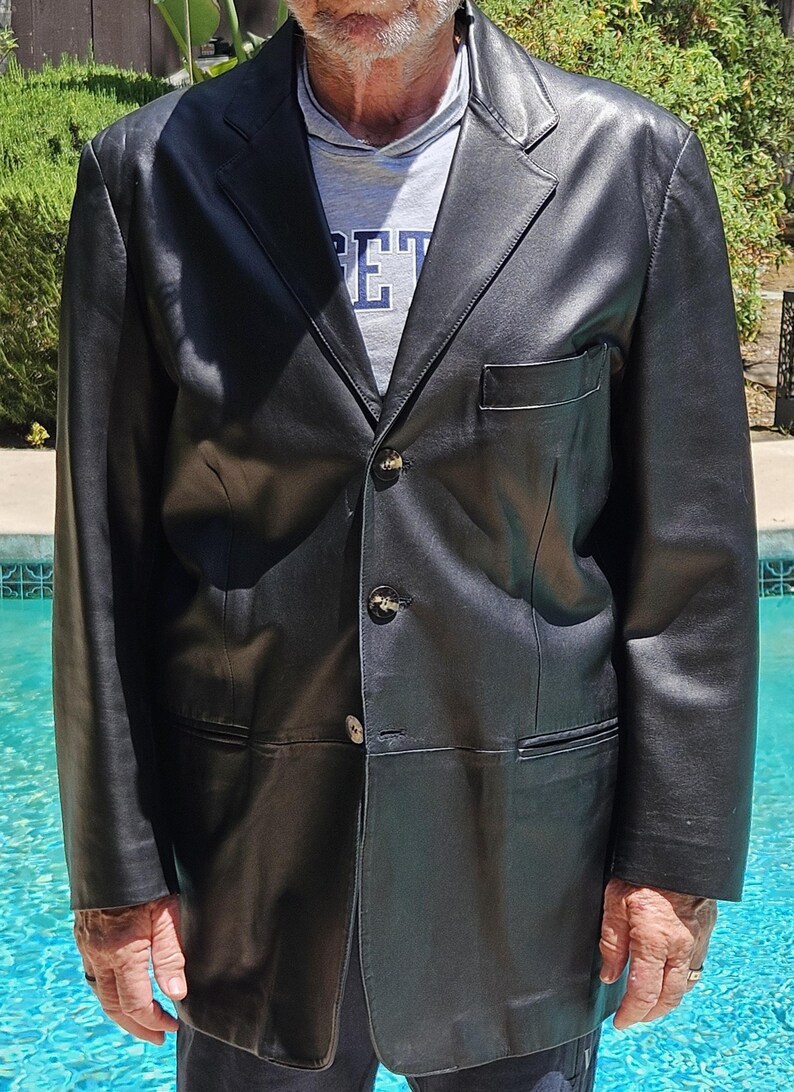 Pu&ograve; includere: Un blazer in pelle nera con revers dentellato, tre bottoni frontali e una tasca sul petto. Il blazer &egrave; indossato sopra una t-shirt grigia con il testo "ET" visibile in lettere blu. Il capo presenta maniche lunghe e un orlo dritto.
