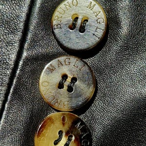 Pu&ograve; includere: Tre bottoni rotondi, marrone chiaro, marmorizzati con venature marrone scuro, fissati su una superficie in pelle nera. Ogni bottone presenta due fori con filo nero e la scritta BRUNO MAGLI impressa in circolo attorno al centro.