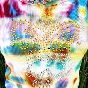 May include: A tie-dye tank top with a skull design made of rhinestones. The tank top features a colorful pattern of blues, purples, yellows, and oranges. A beaded necklace with iridescent and purple beads is around the neckline.