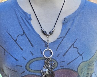 May include: A silver charm necklace with a black cord. The necklace features a heart, elephant, cross, and hand charms. The model is wearing a blue t-shirt with a heart and the word "PEACE".