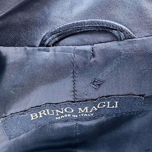 Pu&ograve; includere: L'interno di un capo di colore blu navy scuro con un'etichetta in tessuto nero che riporta il testo ricamato in oro BRUNO MAGLI MADE IN ITALY. L'etichetta &egrave; cucita su una fodera scura e testurizzata con una cucitura curva visibile sopra.