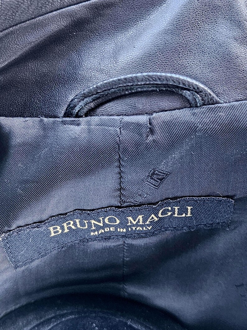 Pu&ograve; includere: L'interno di un capo di colore blu navy scuro con un'etichetta in tessuto nero che riporta il testo ricamato in oro BRUNO MAGLI MADE IN ITALY. L'etichetta &egrave; cucita su una fodera scura e testurizzata con una cucitura curva visibile sopra.