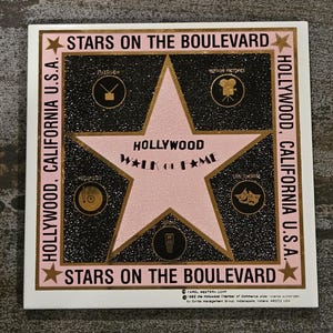 May include: A pink star with a gold outline on a black background. The star has the words "Hollywood Walk of Fame" inscribed on it. The background is surrounded by a black border with the words "Stars on the Boulevard" at the top and bottom. The words "Hollywood, California U.S.A." are on the left and right sides of the border.