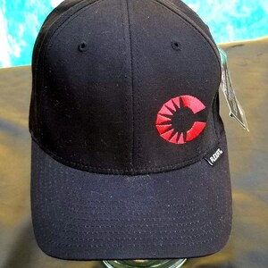 May include: A black baseball cap featuring a red embroidered circular logo on the front panel. The logo consists of a stylized letter C with radiating lines. A small black tag with the white text FLEXFIT is attached to the side of the cap.