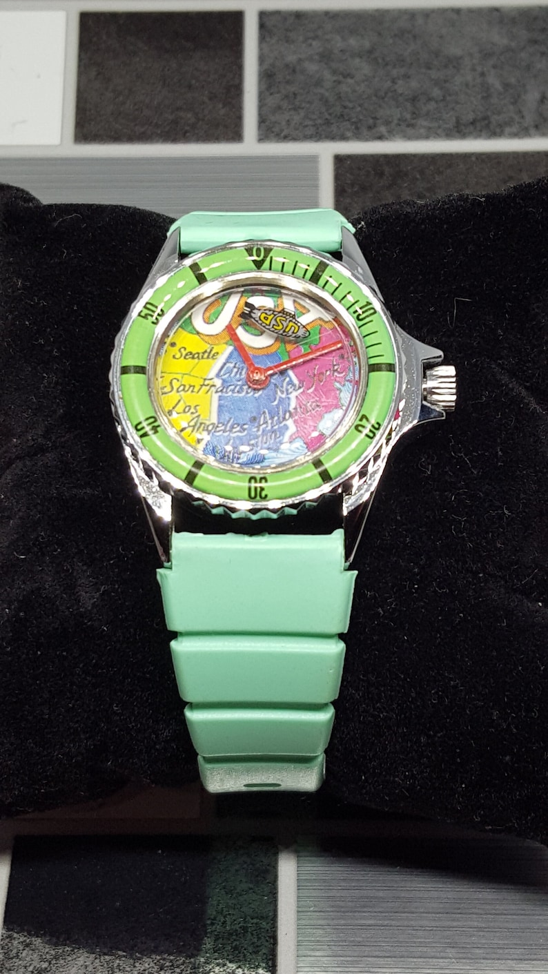 May include: A green wristwatch with a silver band and a map of the United States on the face. The watch face also has the text "USPS" on it.