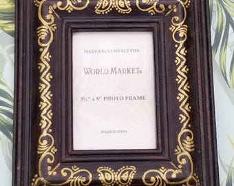 May include: A dark brown wooden picture frame with gold painted details. The frame is decorated with a paisley pattern and has a 3 1/2 inch by 5 inch opening. The frame is made in India and is made exclusively for World Market.