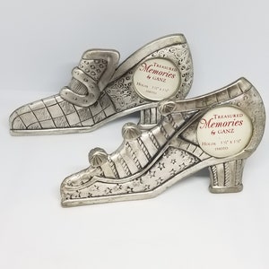 May include: Two silver metal picture frames shaped like shoes. The frames are decorated with stars and stripes. The frames are designed to hold 1 1/2 inch by 1 1/2 inch photos. The frames are labeled "Treasured Memories by Ganz".