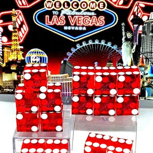 May include: Red dice with white dots are stacked on clear plastic stands. The background features a neon sign that says "Welcome to Fabulous Las Vegas Nevada" with images of the Eiffel Tower, the Statue of Liberty, and a Ferris wheel.