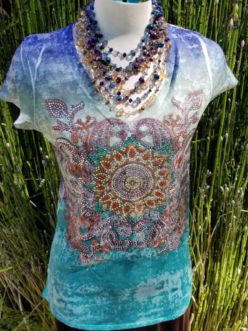 May include: A turquoise blue, white, and purple short-sleeved shirt with a colorful rhinestone design. The shirt has a V-neck and a distressed look.
