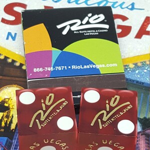 May include: Two red dice with white dots and gold lettering. The dice are from the Rio All-Suite Hotel & Casino in Las Vegas, Nevada.