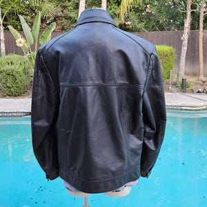 Pu&ograve; includere: Una giacca in pelle nera vista da dietro, caratterizzata da un classico colletto ripiegato, maniche lunghe con polsini abbottonati e una cucitura orizzontale sulla parte superiore della schiena. La giacca ha una finitura liscia e cuciture visibili.