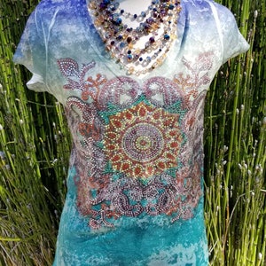 May include: A turquoise blue, white, and purple ombre t-shirt with a large, sparkly, multi-colored mandala design. The shirt has a v-neck and short sleeves.