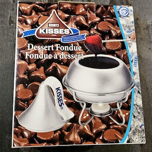 May include: Hershey's Kisses Dessert Fondue box. The box features a silver fondue pot with a strawberry dipped in chocolate. The background is filled with chocolate kisses. The box reads "Dessert Fondue" and "Milk Chocolate".