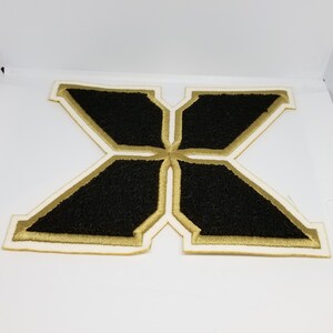 May include: Black felt letter X with a gold outline and white backing. The letter is a patch or applique.