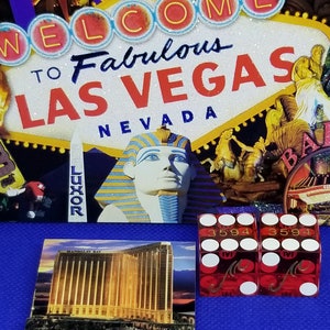 May include: Two red dice with gold lettering and white dots. The dice read "3594" and have the letter "M" on the side. The dice are on a blue surface in front of a postcard of the Mandalay Bay hotel in Las Vegas.